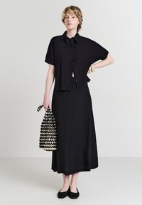 Donna con una camicia nera a maniche corte e una gonna lunga, che tiene una borsa intrecciata bianca e nera, indossando scarpe basse nere e bracciali beige.