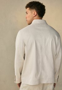 Lichtbeige jas van gestructureerde stof met eenstaande kraag en lange mouwen met knoopmanchetten, die een schone, minimalistische uitstraling laat zien.