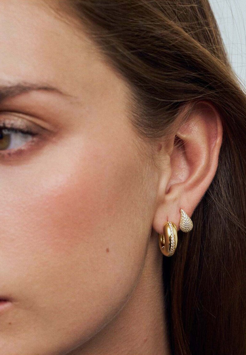 Guldögonringar med en strukturerad hängdesign, som har en liten rad av klara stenar. Bakgrunden är en närbild av ett ansikte med brunt hår.