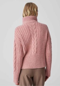 Personne vue de dos portant un pull à col roulé épais en maille torsadée rose avec un motif côtelé et un pantalon marron.