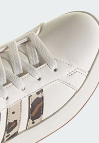 Bijele kožne tenisice s teksturiranim završetkom, s kremastim vezicama i tri smeđe-crne prugaste uzorke sa strane. Dizajn sa zaobljenim vrhom.