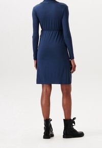 Laivansinis long-sleeve mekko, jossa on tiukka vyötärö, ulottuu juuri polven ylle, valmistettu sileästä kankaasta. Yhdistetty mustiin nauhakengiin nilkkojen ylle.