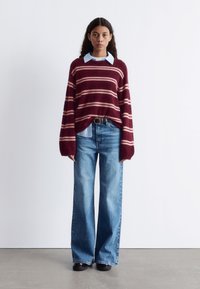 Weinrot gestreifter Pullover aus weichem Material, getragen über einem hellblauen Hemd mit Kragen, kombiniert mit weiten blauen Jeans und schwarzen Schuhen.