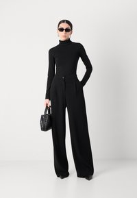 Maglione nero a maniche lunghe con collo alto abbinato a pantaloni neri a gamba larga. Gli accessori includono una borsa strutturata nera e occhiali da sole rettangolari.