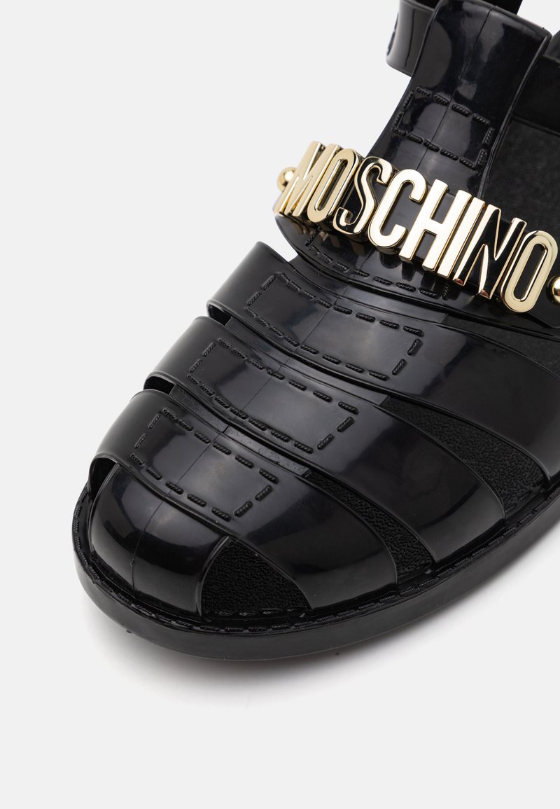 Sandale en cuir verni noir avec des brides croisées, dotée du logo doré "MOSCHINO" et d'un tissu texturé contrastant sur la semelle intérieure.