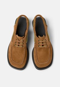 Zapatos de ante marrón de fácil poner con punta redondeada, acentos verdes y suelas de goma negras. Presenta un diseño de doble cordón y un corte en la parte trasera.