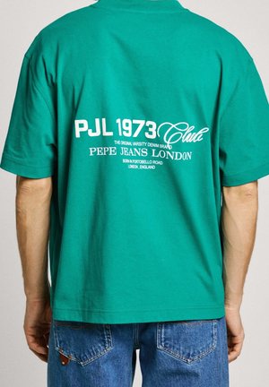 Osoba ubrana w zieloną koszulkę polo z napisem „PJL 1973 Club Pepe Jeans London” na plecach, w połączeniu z niebieskimi dżinsami.