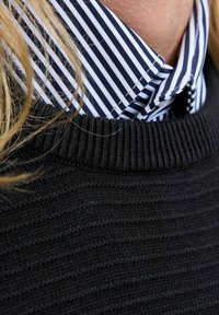 Fekete bordázott pulóver gallérja egy csíkos kék és fehér ing fölött. A hangsúly a kötött anyag textúráján és az ing éles vonalain van.