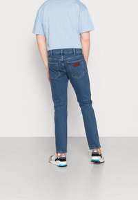 Ljusblå t-shirt, mörkblå slim-fit jeans med fem fickor, brunt läderdetalj på baksidan och grå sneakers med orangea detaljer.