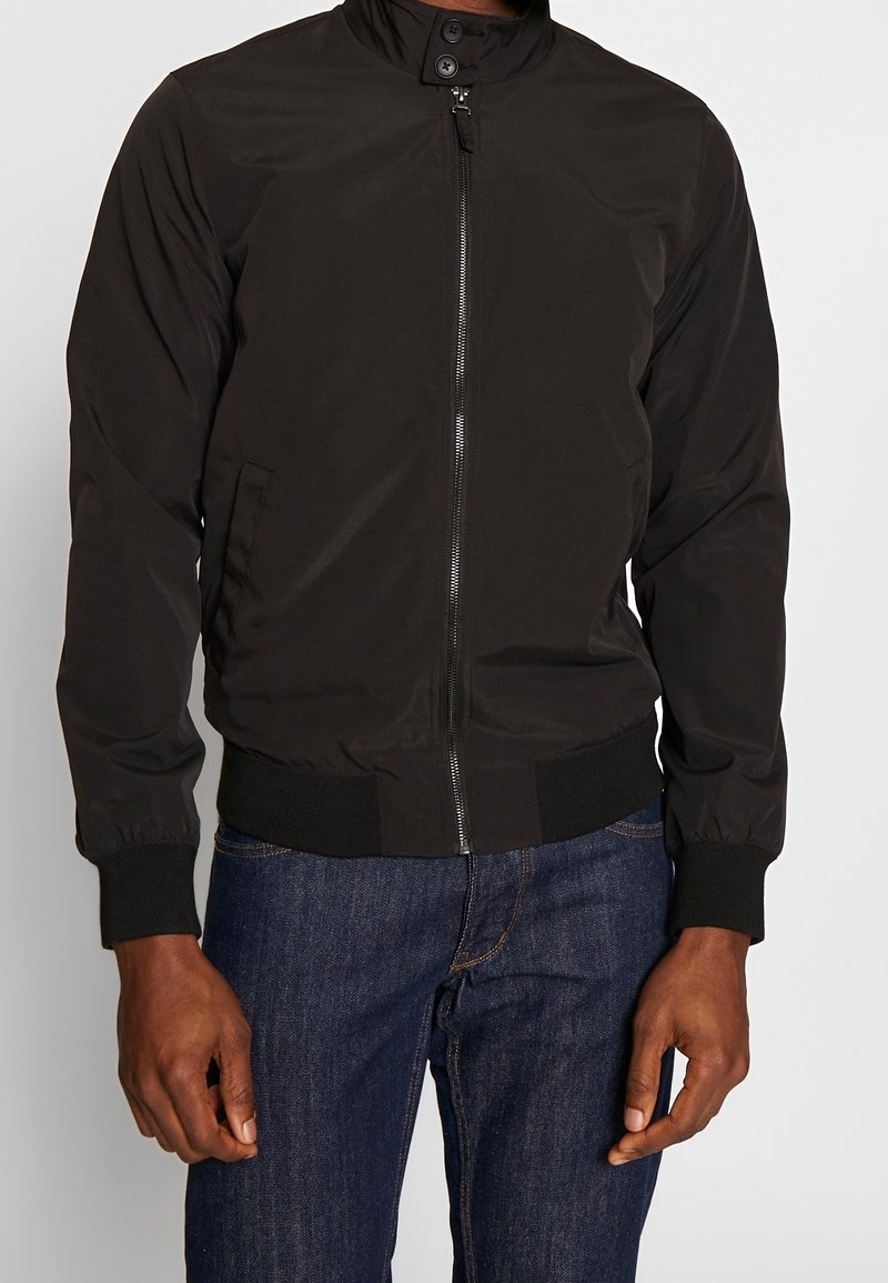 Homme portant une veste bomber noire zippée avec col à boutons et un jean bleu foncé, debout devant un fond clair uni.