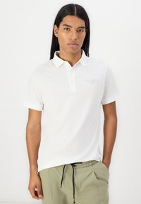 White polo shirt in a soft cotton blend with a classic collar, short sleeves, and subtle logo embroidery on the chest. Paired with green pants.