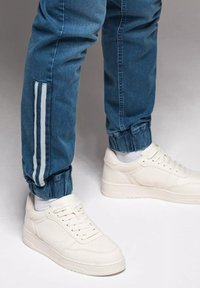 Jasnoniebieskie dżinsowe joggery z mankietem i białymi paskami po bokach, w połączeniu z białymi trampkami o gładkiej fakturze i sznurowanym wzorze.