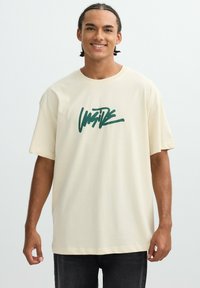 Camiseta de algodón crema con mangas cortas y un corte holgado, que presenta un diseño gráfico en verde en la parte delantera. Jeans oscuros completan el conjunto.