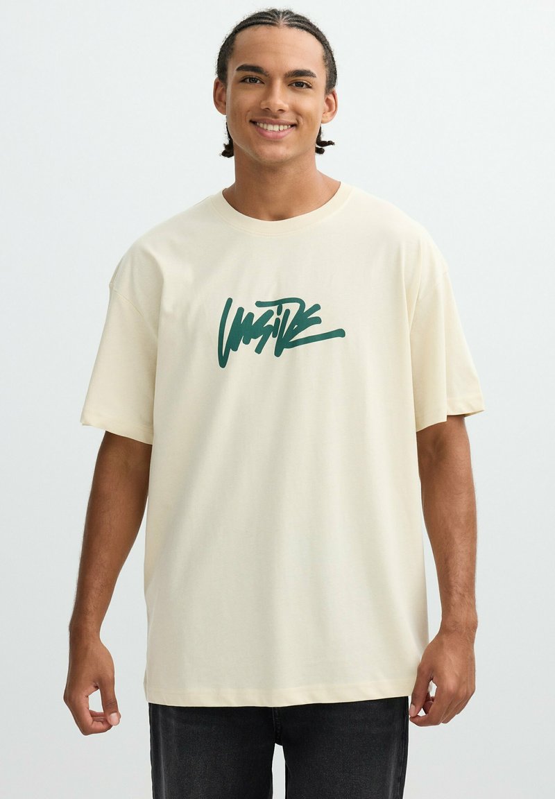 Camiseta de algodón crema con mangas cortas y un corte holgado, que presenta un diseño gráfico en verde en la parte delantera. Jeans oscuros completan el conjunto.