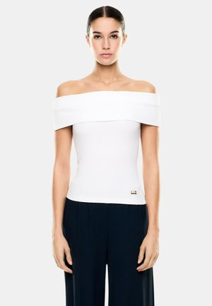 Donna con capelli scuri raccolti, che indossa una maglietta bianca con spalle scoperte e pantaloni larghi scuri, in piedi davanti a uno sfondo chiaro e semplice.