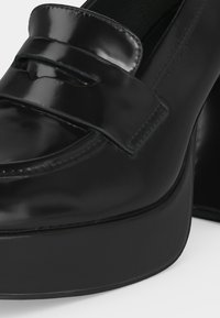 Γυαλιστερό μαύρο δερμάτινο loafers με πλατφόρμα, χοντρό ψηλό τακούνι και κλασική λεπτομέρεια πορτοφολιού στο πάνω εμπρόσθιο μέρος.