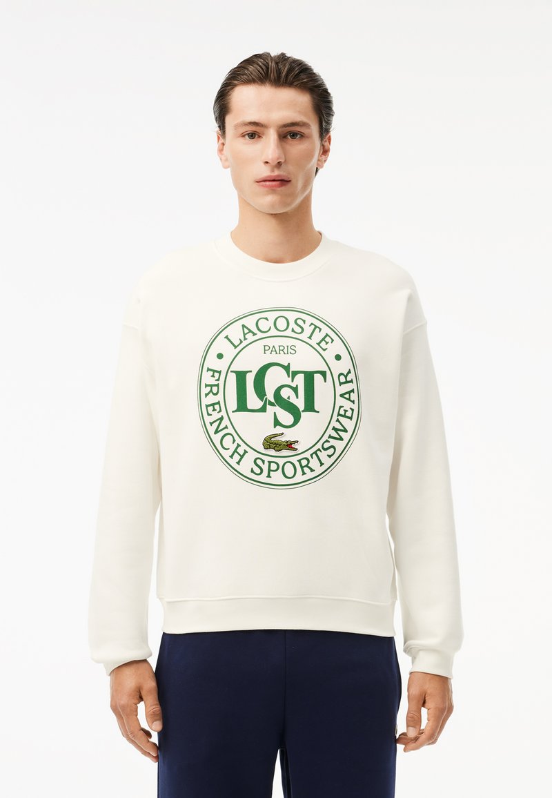 Felpa in cotone crema con grande logo verde con la scritta "LACOSTE," "PARIS," e "FRENCH SPORTSWEAR," colletto rotondo e polsini a costine.