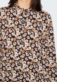 Blusa preta com padrão floral, colarinho com volante e fecho de botão. Apresenta padrões florais em rosa, amarelo e verde em tecido suave.