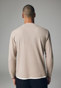 Top beige de manga larga con textura y patrón de canalé. Cuenta con un cuello redondo y puños ajustados, superpuesto a una camisa blanca.