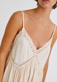 Cremefarbenes Kleid mit dünnen Trägern, ausgestattet mit einer Spitzenkante am Ausschnitt und gefaltetem Stoff für Textur. Einfaches und leichtes Design.
