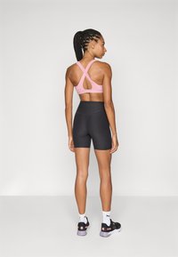 Rosa Sport-BH mit überkreuztem Rückendesign, kombiniert mit schwarzen Shorts mit hoher Taille. Das Model trägt schwarze Sportschuhe und weiße Socken.