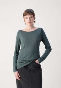 Žalia ilgų rankovių palaidinė, pagaminta iš tekstūruotos medžiagos, su plačia kaklo iškirpte, derinama su juoda džinsine sijonu.