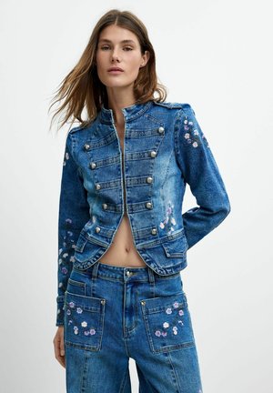 Donna che indossa una giacca di denim blu coordinata e jeans con ricamo floreale e bottoni argentati su uno sfondo chiaro semplice.