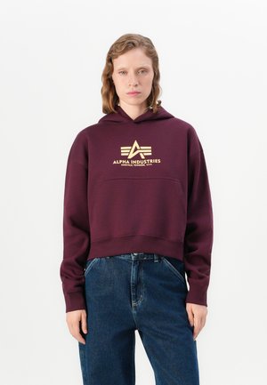 Burgunderfarget, kort hoodie med gull "Alpha Industries" logo. Har en frontlomme og justerbar hette med snøring. Glatt tekstur.