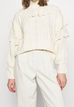 Kobieta ubrana w kremowy, gruby sweter w warkocze z koronkowymi falbankami na ramionach oraz kremowe spodnie z wysokim stanem na białym tle.