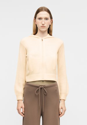 Sirge juustega naine, kellel on seljas kreemikas lukuga kapuutsiga pusa ja pruunid nööriga püksid, seisab ühetaolise heleda tausta ees.