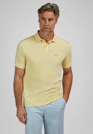 Mann mit grauen Haaren und Bart, der ein hellgelbes Poloshirt und hellblaue Hosen trägt, mit einer Hand in der Tasche stehend vor einem schlichten Hintergrund.