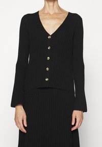 Černý žebrovaný kardigan má V-výstřih, dlouhé rukávy a sedm kulatých kontrastních knoflíků vpředu pro zapínání.