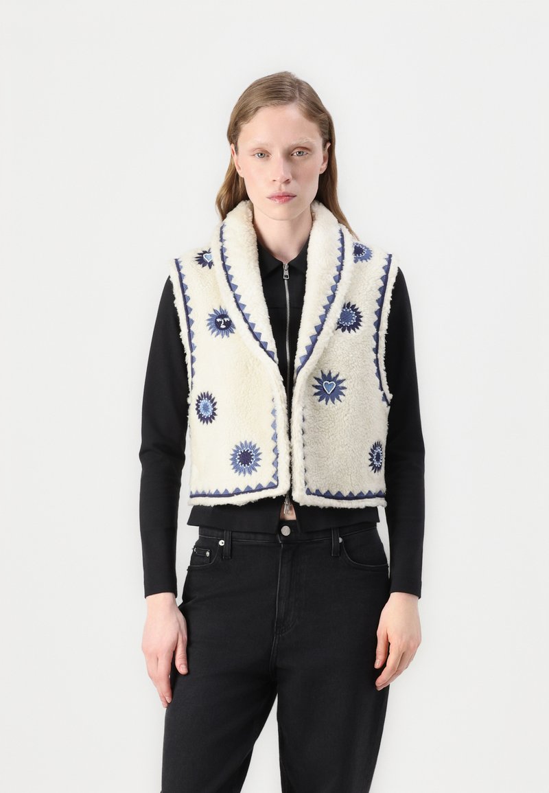 Fleeceväst i krämfärg med blå broderade solmotiv, bred krage och vågig trim; bärs över en svart topp och jeans.