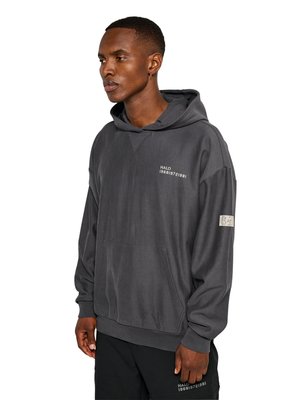 Man wearing dark gray hoodie and black pants, standing sideways with hands relaxed, hoodie has small white text on chest and sleeve.