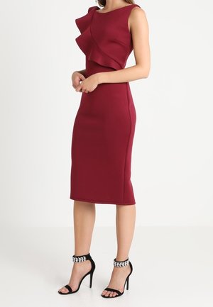 Robe bordeaux sans manches avec un col asymétrique à volants ; silhouette ajustée et longueur au genou. Portée avec des sandales à talons noirs.