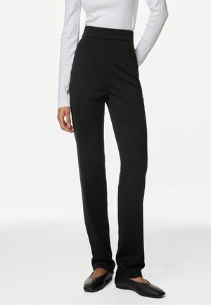 Pantalons noirs taille haute en tissu lisse, avec une coupe ajustée et des jambes droites. Portés avec un haut ajusté à manches longues blanc.