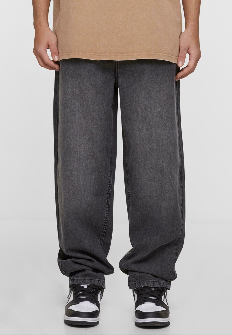 Jeans de pierna ancha en gris oscuro con una textura desvaída, con costuras laterales y tobillos remangados, combinados con zapatillas negras y blancas.