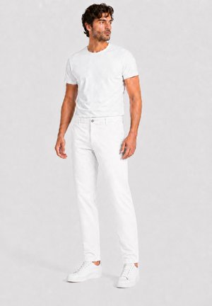 Man met krullend haar, gekleed in een nauwsluitend wit T-shirt, witte spijkerbroek en witte sneakers, staat en kijkt naar de zijkant.