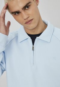 Jeune personne aux cheveux blonds courts ajustant sa tête, portant un sweat à fermeture éclair bleu clair par-dessus un t-shirt noir, sur fond neutre.