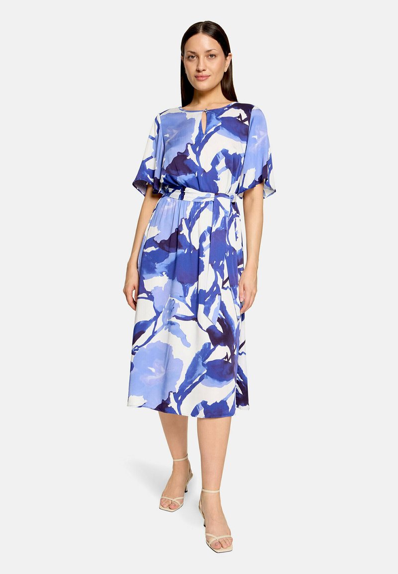 Femme portant une robe fleurie abstraite bleu et blanc longueur genoux, à manches courtes et ceinture nouée à la taille, associée à des talons beiges à brides.