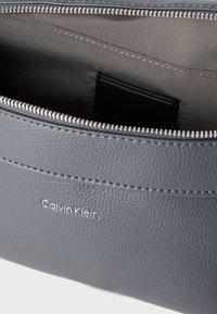 Γκρι δερμάτινο θήκη με φερμουάρ, με ομαλή υφή και ανάγλυφο λογότυπο "Calvin Klein" στο μπροστινό μέρος. Το εσωτερικό είναι επενδεδυμένο με ύφασμα.