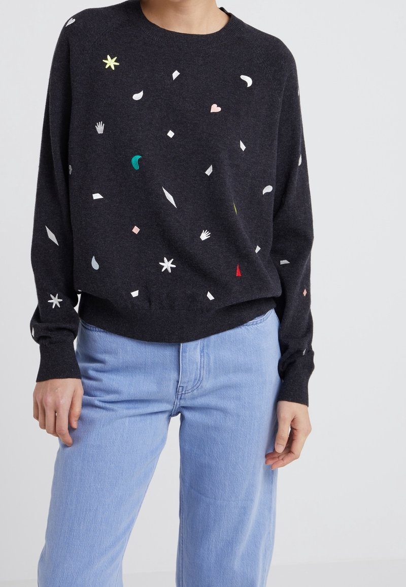 Pull gris foncé avec des formes abstraites colorées comprenant des mains, des cœurs et des étoiles, porté avec un jean bleu clair par une personne debout.