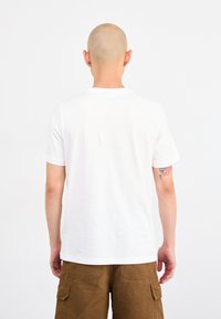 Person med barbert hode iført en enkel hvit t-skjorte og brune cargo-shorts, som viser en fargerik tatovering på venstre arm.
