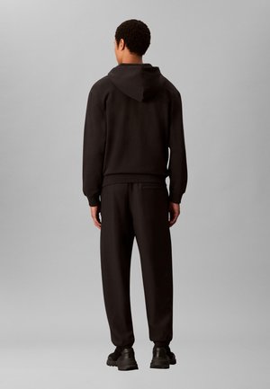 Homme de dos portant un sweat à capuche noir, un pantalon de survêtement noir assorti et des baskets noires épaisses, devant un fond gris uni.