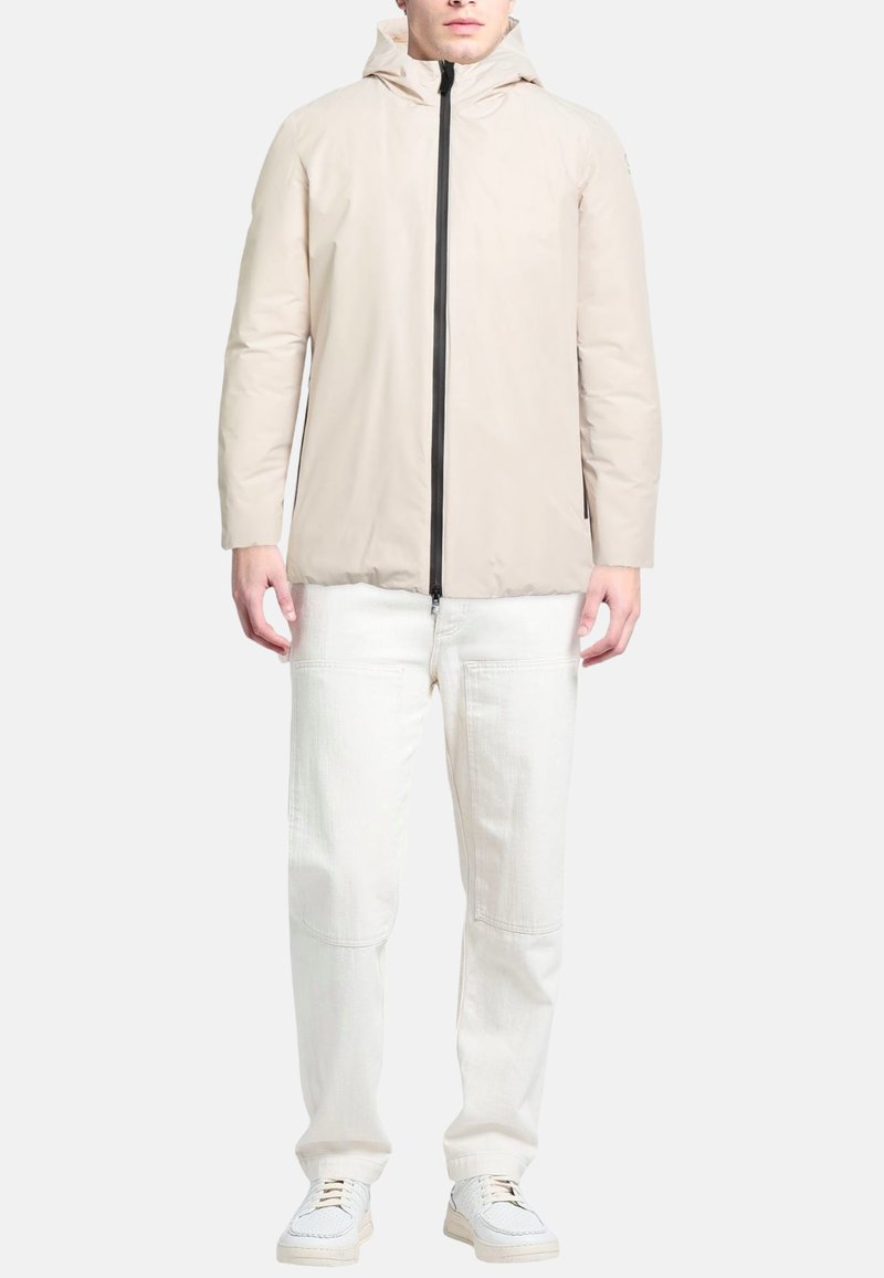 Lichtbeige jas met capuchon, voorzien van een ritssluiting aan de voorkant, lange mouwen en een gladde textuur. Gecombineerd met een witte broek en sneakers.