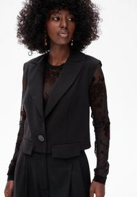 Czarna, dopasowana kamizelka z wyraźnymi ramionami, z zapięciem na jeden guzik i dwoma przednimi kieszeniami. Pod nią znajduje się przezroczysta czarna bluzka z haftem w kwiaty.