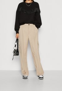 Sudadera negra con volantes en capas y detalles brillantes, pantalones cargo beige con bolsillos frontales, bolso negro y zapatillas negras y blancas.