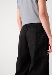 Schwarze Cargo-Hosen mit elastischem Bund, seitlichen Taschen und einer aufgesetzten Gesäßtasche. Der Stoff hat eine glatte Textur und ein mattes Finish.