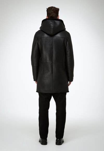 Schwarzer Ledermantel mit Kapuze, der eine glatte Textur, lange Länge und ein weiches Futter entlang des Kapuzenrands aufweist. Über schwarzen Hosen getragen.