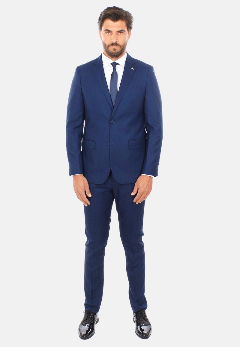 Completo blu navy con una vestibilità slim, giacca monopetto, pantaloni abbinati e una camicia bianca con una cravatta scura. Scarpe lucide nere.